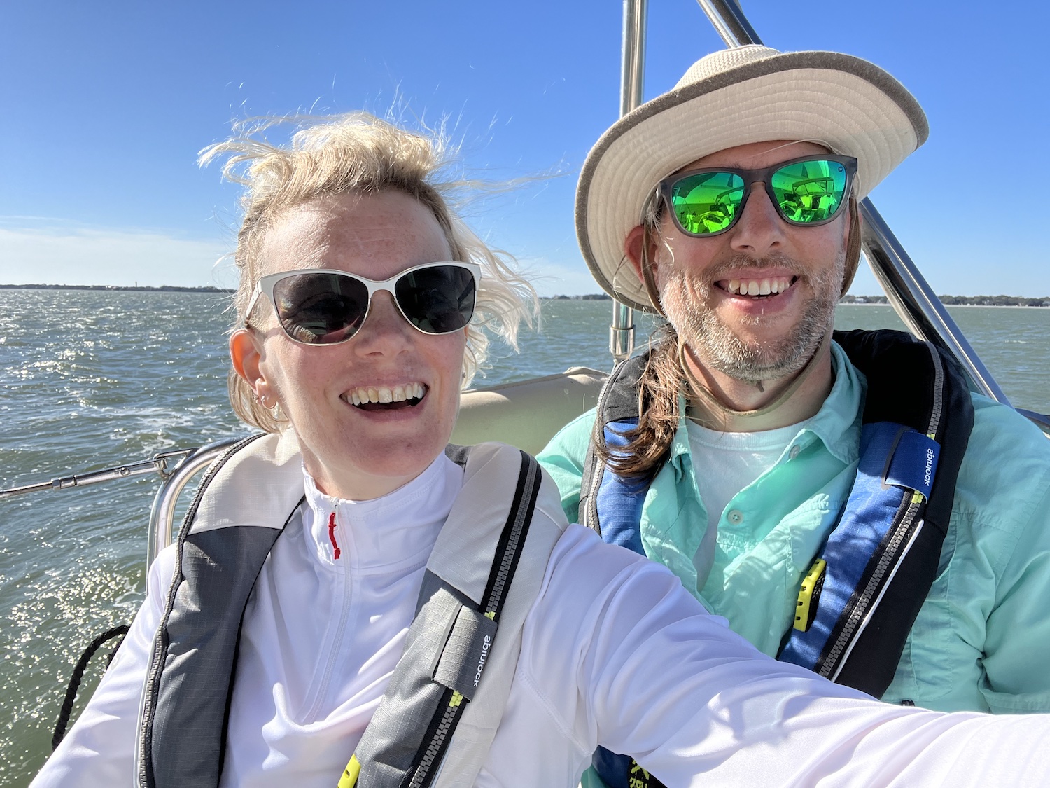 pinky has blonde short hair, pulled back and is wearing a while long sleeve shirt with a lifejacket and sunglasses on. charles has long hair pulled in a pony tail, wearing sunglasses and a hat. he's also wearing a long sleeve teal shirt and a life jacket. the ocean is in the background as they are on a sailboat