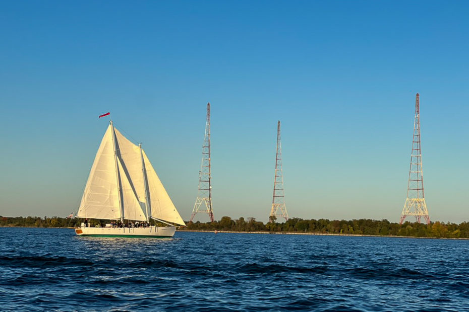 schooner woodwind, a large multi-mast sailboat, sailing across blue water with full sails out and people out on deck enjoying the sunset. three radio towers are in the background on land.
