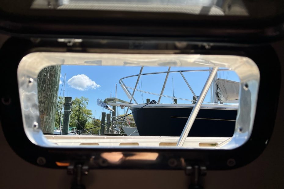 viewing the outside through the frame of a portlight. there is a small white cloud floating in a deep blue sky. the bow of a large boat is in view as well as trees in the background.