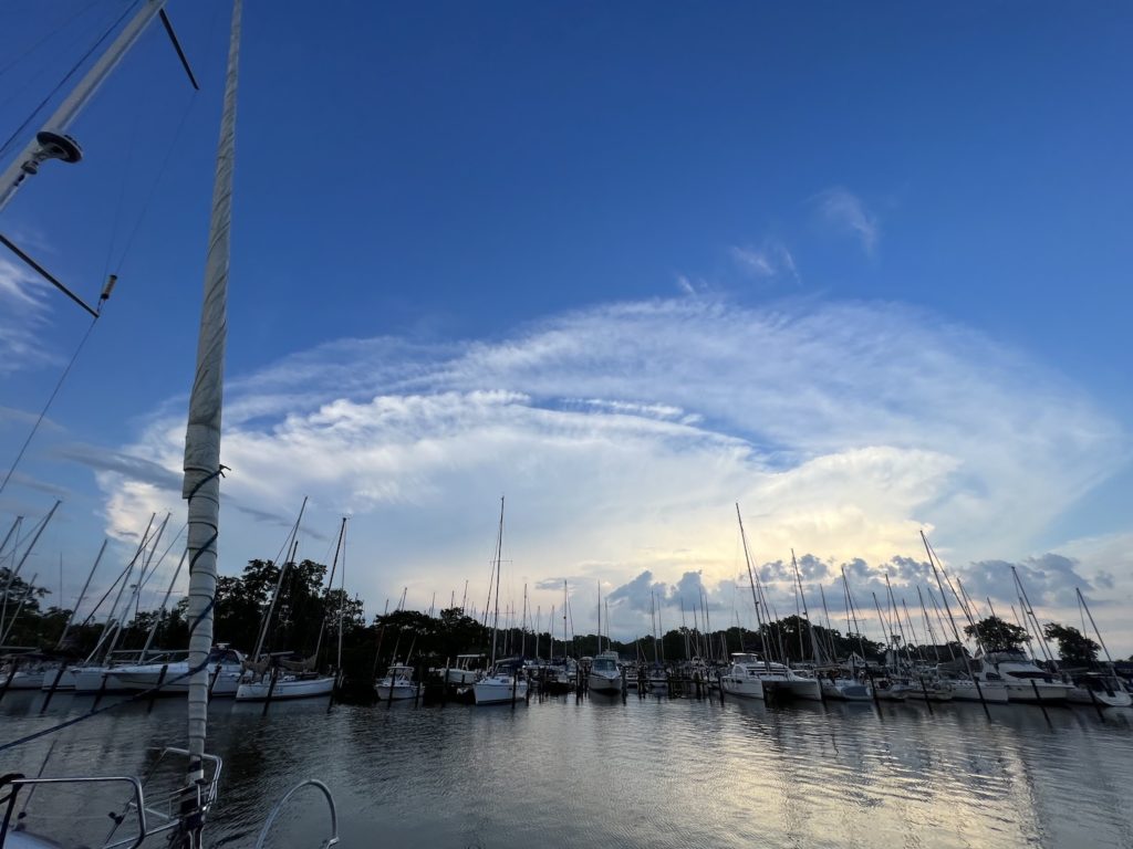 wide angle view of a big blue sky with big storm clouds in the distance. the white clouds are the top of a huge storm system that's moving east, and has the shape of a mushroom cloud. there are boats in the water at a marina in the foreground.
