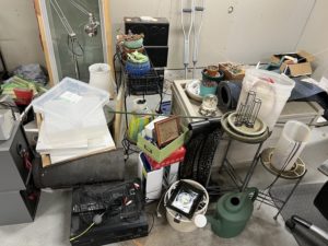 a pile of random stuff in a garage. items include picture frames, a snow globe, crutches, house windows, plant stands, plant pots, plastic containers, old stereo, watering can, lamps, rugs, mats, and more.