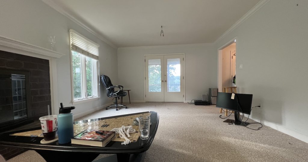 wide angle view of a bare living room. there's an empty fireplace to the left. a coffee table is in the foreground with a couple of drinks on it. there's a monitor on the floor to the right. a lonely office chair sits in the back corner. the walls are bare.
