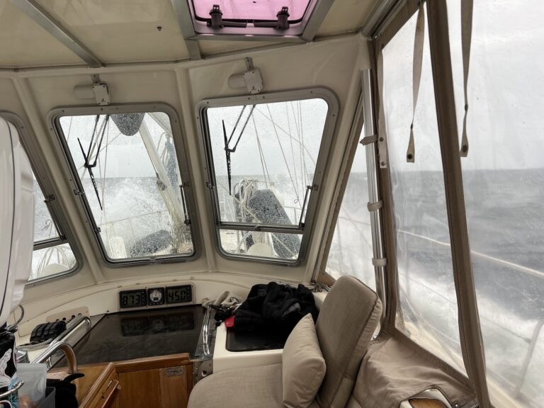 inside the cockpit of a sailboat with grey sea water splashing over the bow of the boat