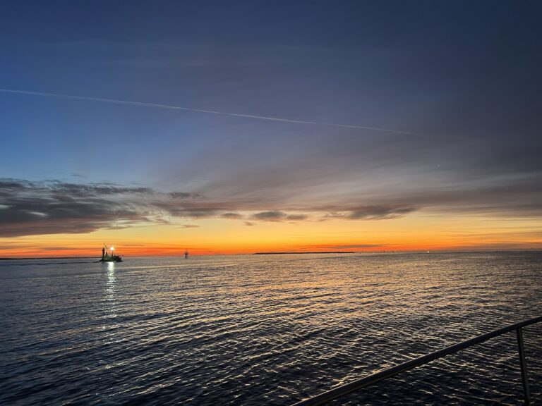 landscape view of big dark sky with a bright yellow/orage/red horizon hinting at the sunrise. a fishing boat with lights on is passing from the left across a dark blue sea