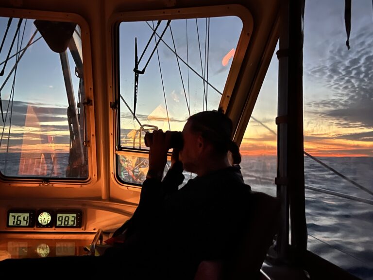 from inside the glowing cockpit, the sunsets over the ocean in the background and charlie is shadowed and looking through binoculars