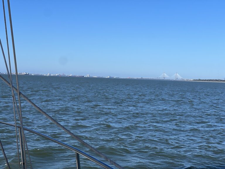 from the boat, a view of charleston harbor under blue skies which is mostly blue water with a large Cooper River bridge in the background along with the Charleston harbor cityscape.