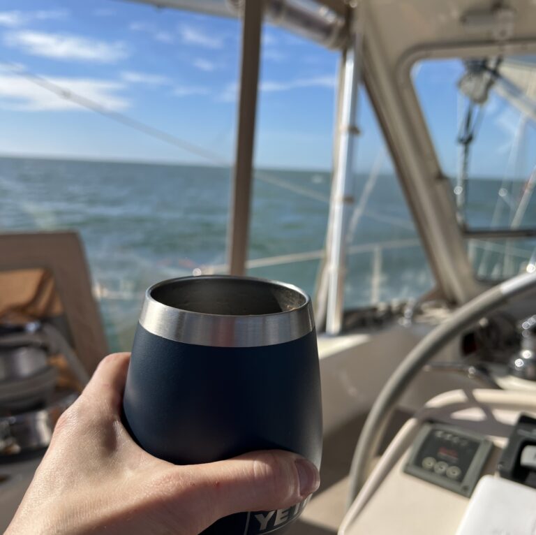 a hand holds an insulated cup of coffee while sitting in the sailboat cockpit with the blue sky, clouds, and ocean in the background