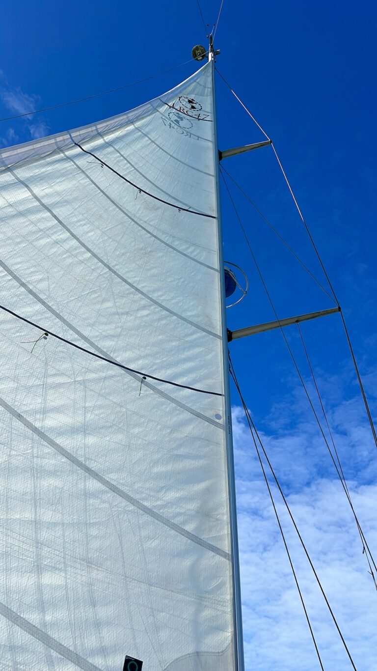 view of the white mainsail fully deployed with a bright blue sky in the background with a few clouds