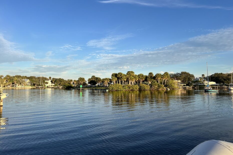 View of a protected Florida river basin surrounded by palm trees with sailboats anchored nearby, and a few houses peeking out from the trees. Blue skies with a few clouds above.
