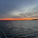 Looking out from the deck of a sailboat, over dark waters towards the horizon where daylight is breaking in an orange and pink sunrise in the overcast sky
