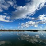 A river of calm flat water with bright blue sky and fluffy white clouds above, reflected in the calm water. A river bank with trees and brown grass cuts horizontally in the middle of the photo.