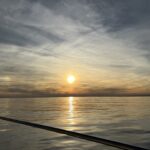 Looking over the railing of a sailboat, the ocean is flat calm with only a few ripples. The yellow, orange sun is setting in the distance with high atmosphere clouds above.