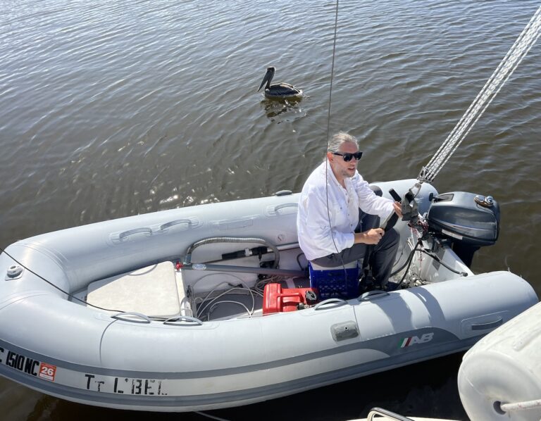 A man in a white sunshirt & sunglasses sites at the back of an inflatable rigid dinghy with an outboard motor on the back. A curious pelican floats in the water nearby, clearly thinking fish may be in his future.