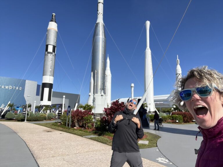 A fun picture of a woman in the foreground smiling an openmouthed smile and a man looking up with a similar openmouthed smile. They are both excited to in visiting the rocket garden. Several rockets are scattered behind them, and a deep blue sky is overhead.