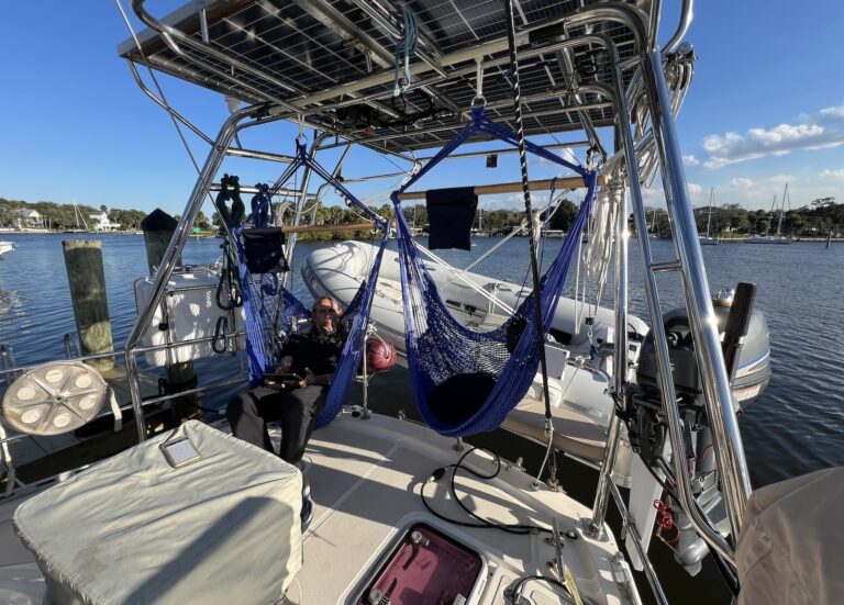 Looking at the back of a sailboat at a large solar arch with 2 net swings hanging from the bars. A man sits in one of them demonstrating how they work. A protected river basin is in the background with calm water, clear blue skies, and anchored sailboats