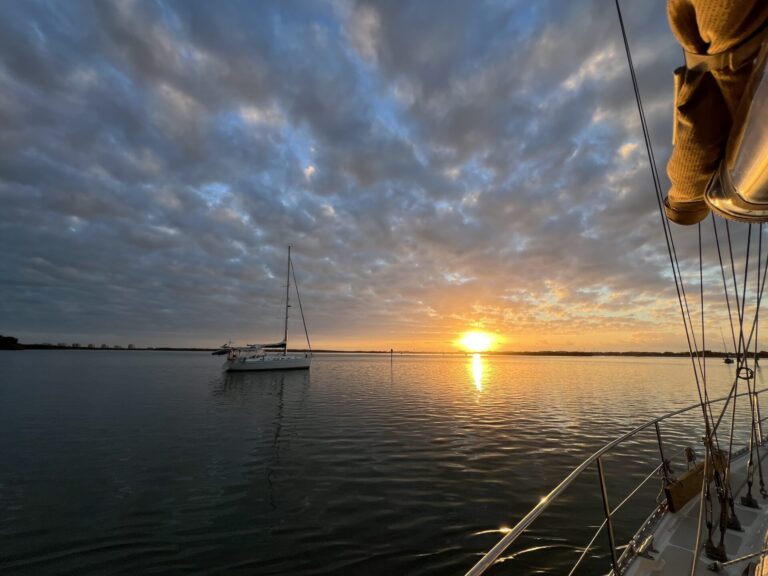 Looking out over the water from the deck of a sailboat, a bright yellow sun rises just above the horizon. The sky is a mix of grey and pink clouds with a bit of blue peeking through. The water is calm, and a sailboat is anchored just to the left of the sunrise.