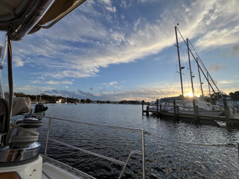 Looking out from a sailboat cockpit at a river basin with a marina dock to the right with several sailboats. The sun sets in bright yellow just beyond those boast. To the left and in the background, there are ripples on the water, sailboats anchored in the distance, and a blue sky with bands of white clouds.