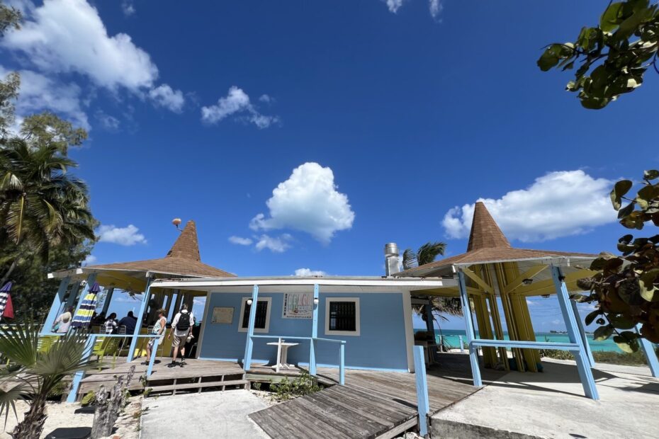 a small blue building is in the center and is flanked by 2 hut like structures with no walls and lots of walkways. it's a restaurant that sits on the beach under clear bright blue skies with a few clouds