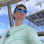 A young woman in sunglasses and a sunshirt looks out from her perch in the cockpit of a sailboat with antennaes and solar panels in the background