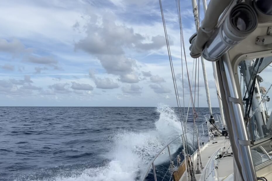 Looking down the port side of a sail boat, with a bow wave cresting off to the left as the boat moves through the blue ocean with a blue sky and white fluffy clouds