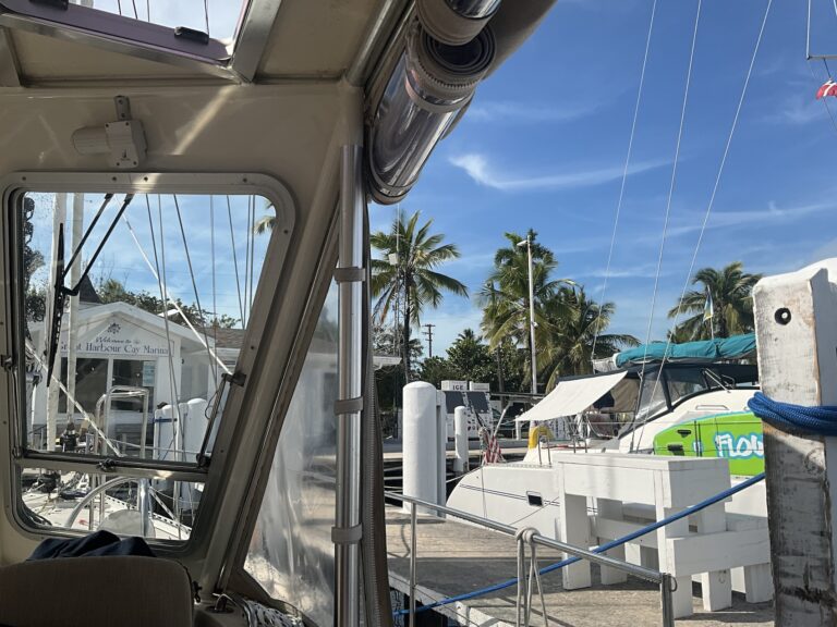 A view from inside the cockpit of a docked sailboat looking out a a marina with other boats and palm trees against a clear blue sky. The sign over the marina office says "Welcome to Great Harbour Cay Marina"