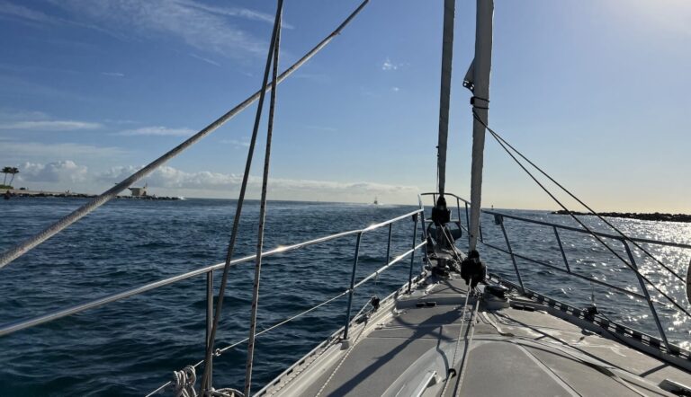 Looking out across the boat of a sailboat with sails furled, blue ocean is calm ahead, as the boat exits an inlet