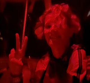 A night shot of Pinky, with wild curley hair and making a peace sign with her fingers. The shot is red hued and she's got a life jacket on.