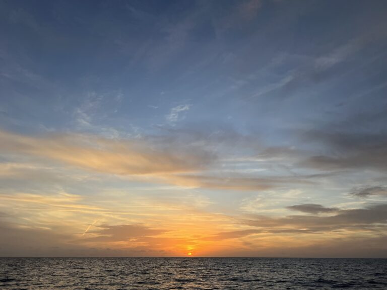 A tiny orange sun sets far across a vast blue ocean with faint clouds in shades of pink and orange as the sky fades to a dark blue
