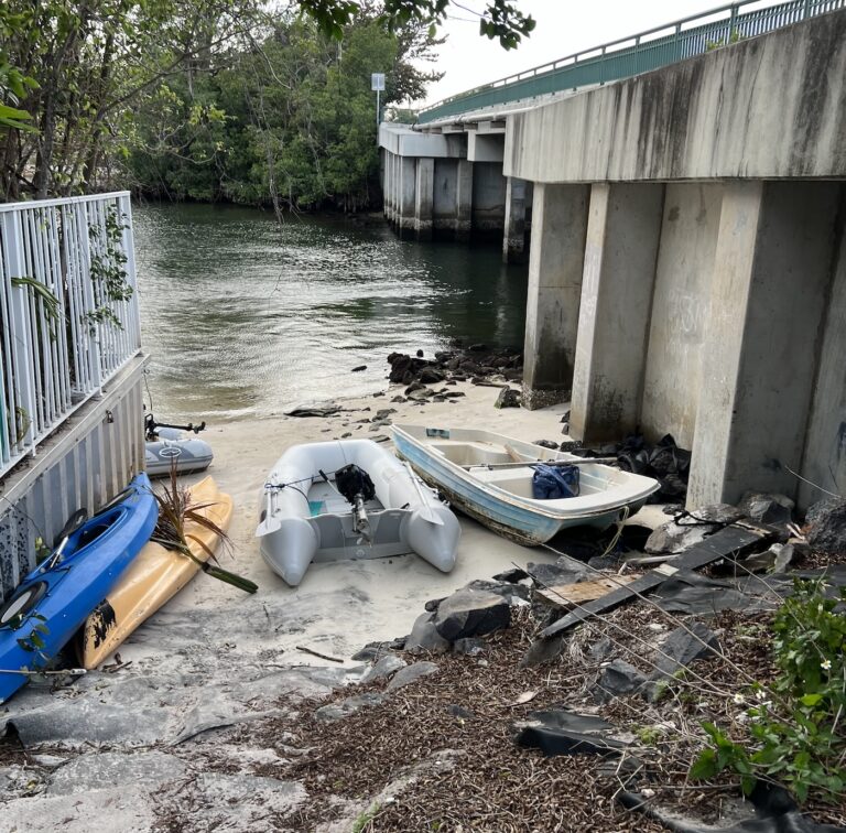 Looking down a narrow beach with kayaks on the left, and 2 inflatable dinghies on the right with a road bridge to the right and a fenced area to the left. A creek is beyond the beach