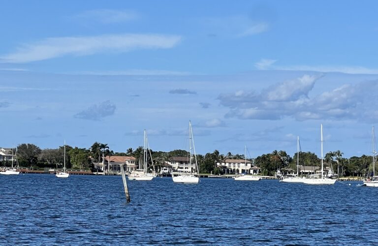 A blue water anchorage with several sailboat anchored out and large houses in the background. A blue sky is above with a few white and grey clouds.