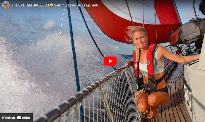 woman in a life jacket kneels on the side deck of a sailboat with the sail in the background and water splashing up on the side of the boat