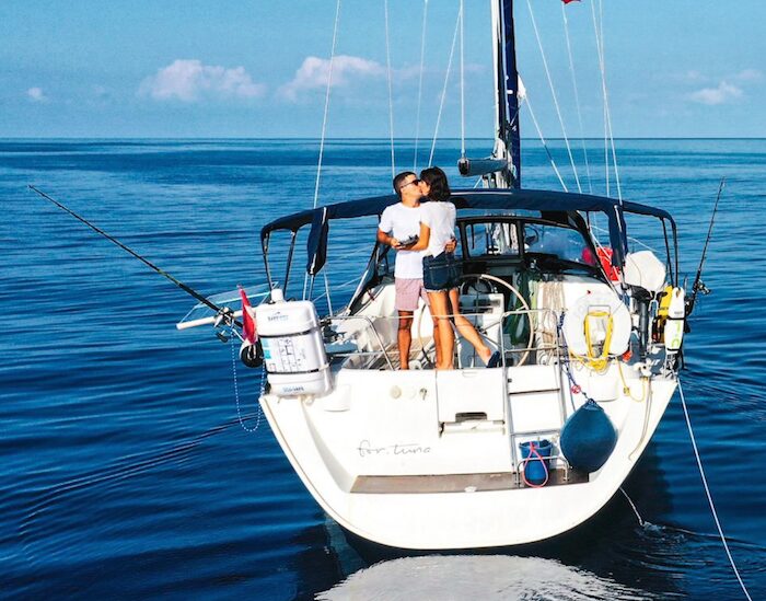 man and woman kissing on the aft deck of the sailboat, which is geared out with fishing rods, a liferaft, fenders, and other typical sailing gear. the sailboat is floating in flat calm deep blue water