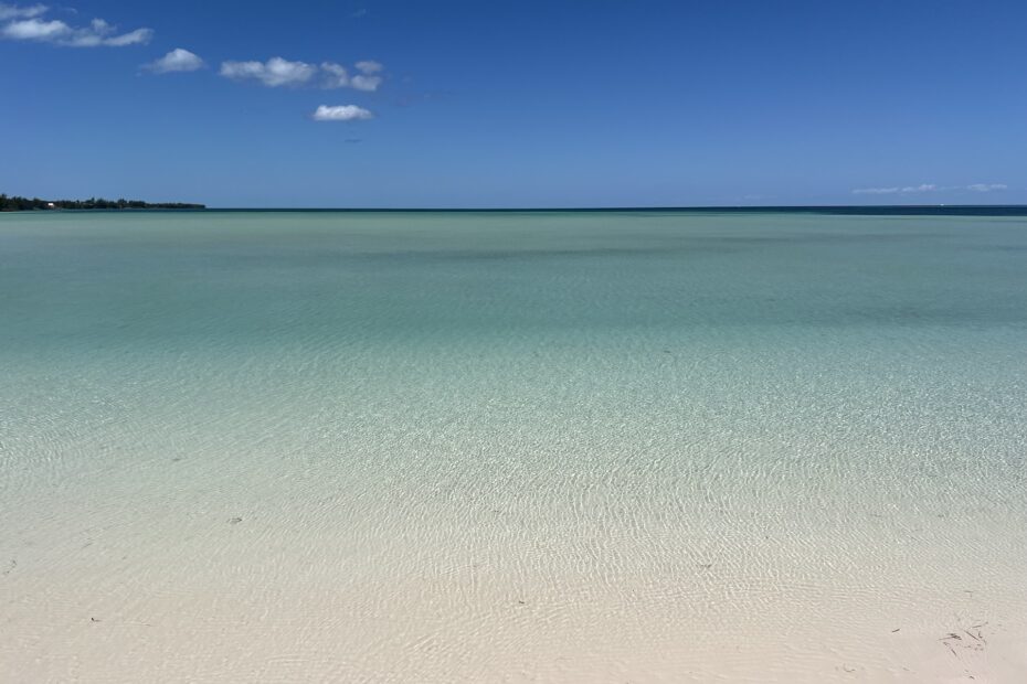 white sandy beach in the foreground that's covered with crystal clear turquoise water extending to the horizon of a bright blue sky with a couple of clouds and land in the far distance