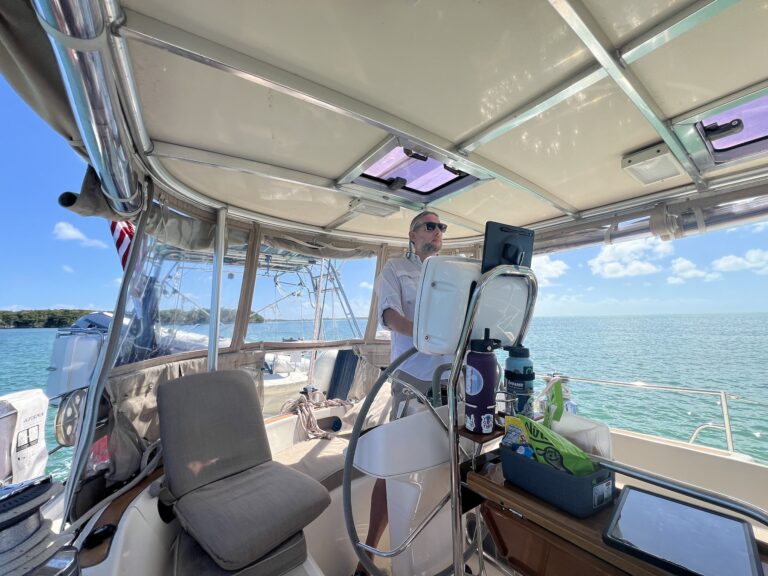 view of a sailboat cockpit looking aft at a man standing at the helm. there's a forward-facing seat to starboard of the helmsman and a snack box and bottles of water on the cockpit table.