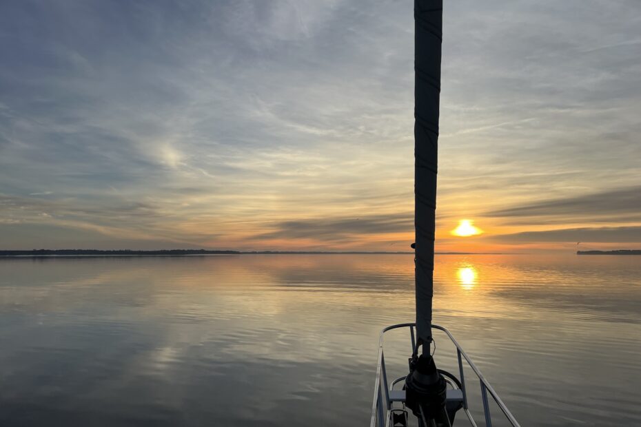 sunrise in a cloud-streaked sky over flat calm water, looking out from the bow of a sailboat with the headsail in view