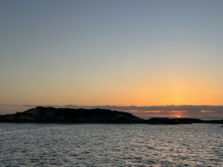 sun just rising over the horizon of a clear sky with an island in the foreground and ocean around it.