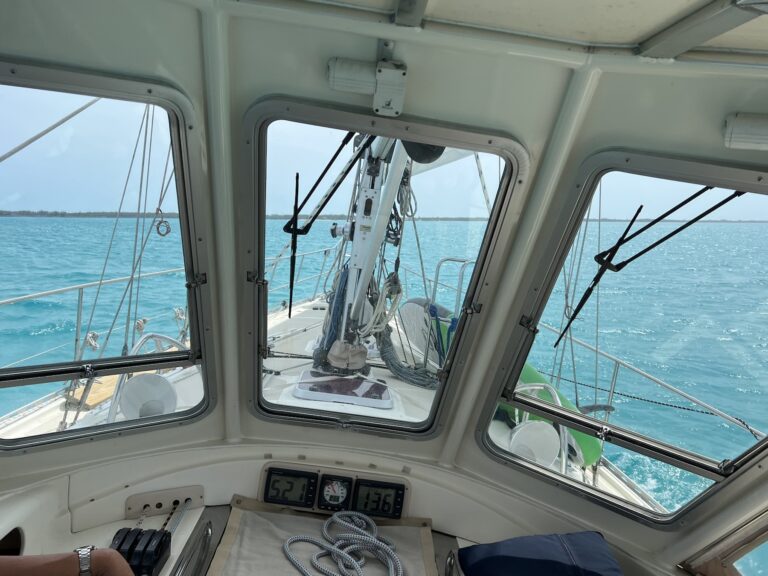 view from inside the sailboat cockpit. looking through 3 big glass windows at turquoise water. sailing instruments just below the windows show speed of 5.2 knots and depth of 13.6 feet. The boat is clearly heeled over 5-10 degrees