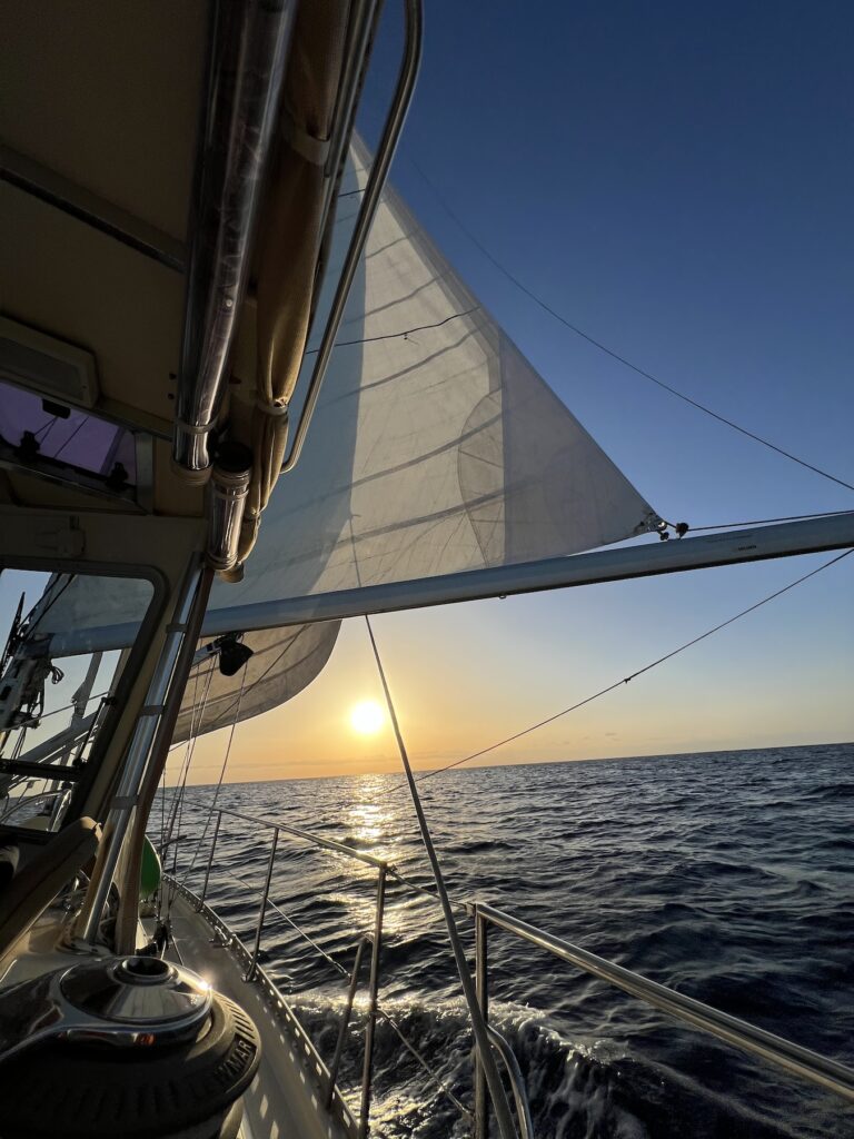 view from the deck of sailboat, the main sail and the genoa unfurled, with a sunrise over the ocean ahead