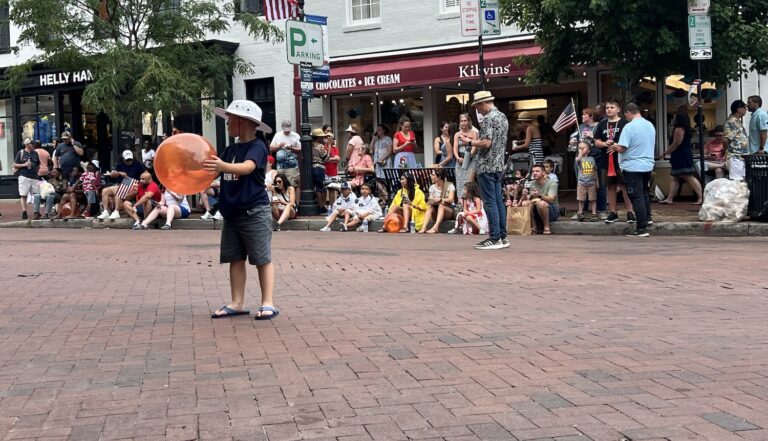 boy standing in the middle of brick-laid street holding an orange beach ball. kids and adults sit and stand on the curb across the street, waiting on a parade. in the background, an ice cream store has people standing in line.