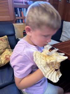 boy sitting inside a boat on a blue settee holding a conch horn to his mouth.