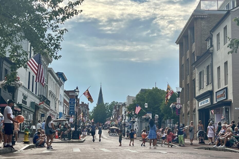 view of main street Annapolis with no cars on it and people walking and scootering down the street. Flags hang on the posts on each side of the street which is lined with businesses. a church steeple pokes up at the end of the street. the sky is blue with some clouds.