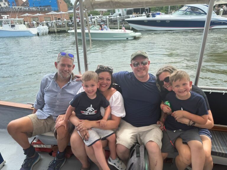 a smiling family sits together on a boat with power boats in the background. there are 2 adult men and 2 adult women who have kids on their laps.