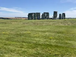 stonehenge, a line of large rocks arranged in a circle, is in the middle of the photo amidst a green grass field and a bright blue summer sky with tourists in the background