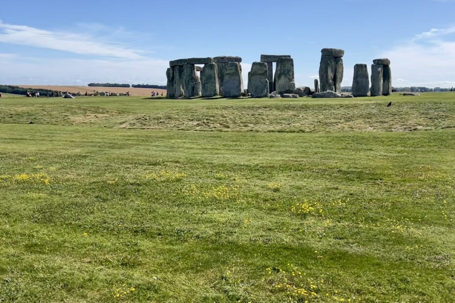 stonehenge, a line of large rocks arranged in a circle, is in the middle of the photo amidst a green grass field and a bright blue summer sky with tourists in the background