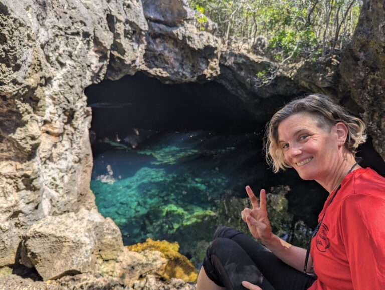 A woman smiles and flashes a peace sign while sitting at the edge of a rocky blue hole or cave pool in Long Island, Bahamas. The sunlight illuminates the crystal-clear water beneath, revealing underwater rocks and vibrant blues.