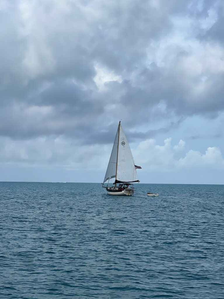 A small sailboat named Mischief sails under partially reefed main on turquoise Bahamian waters, with a wooden dinghy in tow and dramatic clouds above.