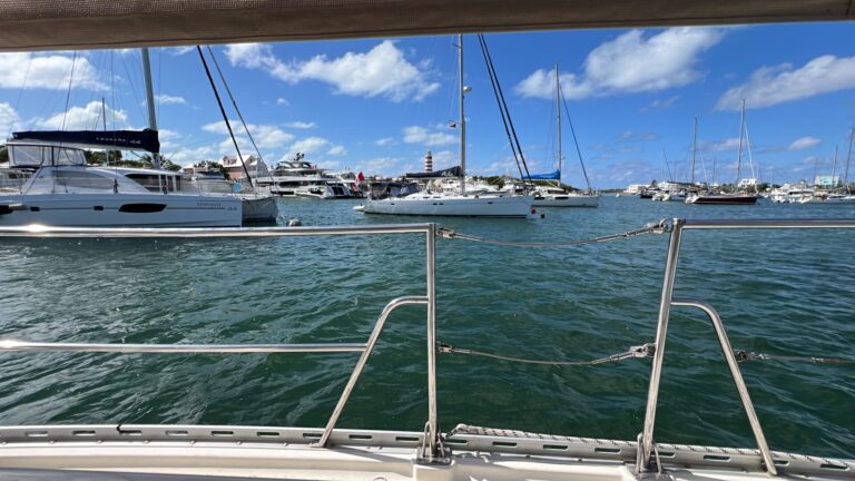 View from a sailboat looking out at a busy mooring field in Hope Town, Bahamas, with catamarans, monohulls, and large motor yachts anchored against a backdrop of pastel buildings and the iconic red-and-white striped Hope Town Lighthouse.
