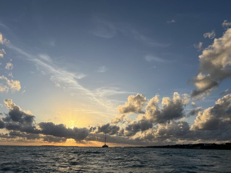 A dramatic sunset over the water near Spanish Wells, Bahamas, with golden rays streaming through low clouds and a solitary sailboat silhouetted against the horizon under a vast sky filled with scattered cumulus clouds.