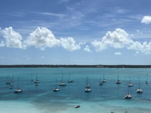 A panoramic view of turquoise water and dozens of anchored sailboats at Stocking Island in the Bahamas under a bright blue sky with fluffy clouds.