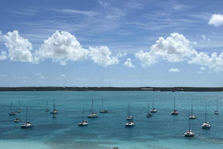 A panoramic view of turquoise water and dozens of anchored sailboats at Stocking Island in the Bahamas under a bright blue sky with fluffy clouds.